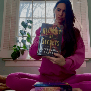 Blog author sitting, holding a copy or Alchemy of Secrets by Stephanie Garber. 